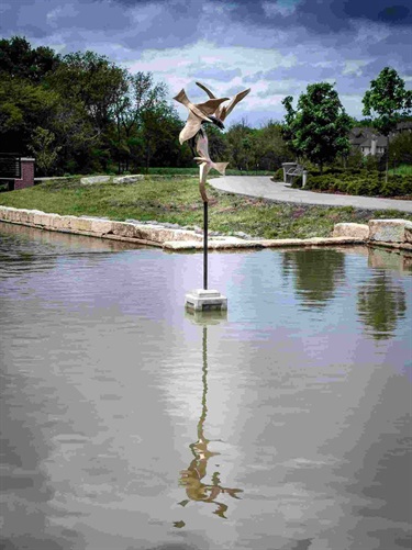 Metal sculpture of birds in the water. Frenzied Flight: Located at Central Green Park, this sculpture by Lenexa artist Robin Richerson, is an abstract depiction of three birds in flight, fighting for the same fish.
