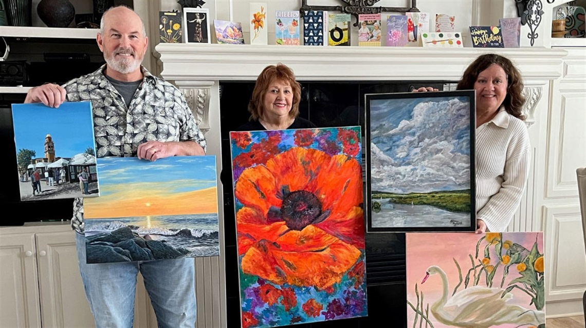 smiling man and two women holding up paintings in front of fireplace