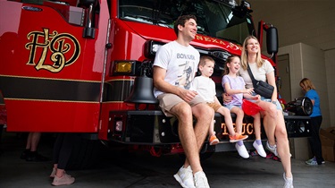 Family with young kids pose seated on front bumper of fire engine