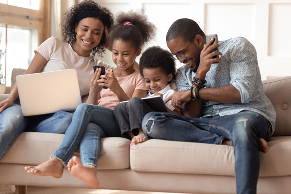 parents and three kids sitting on couch using laptop, tablet, cell phone