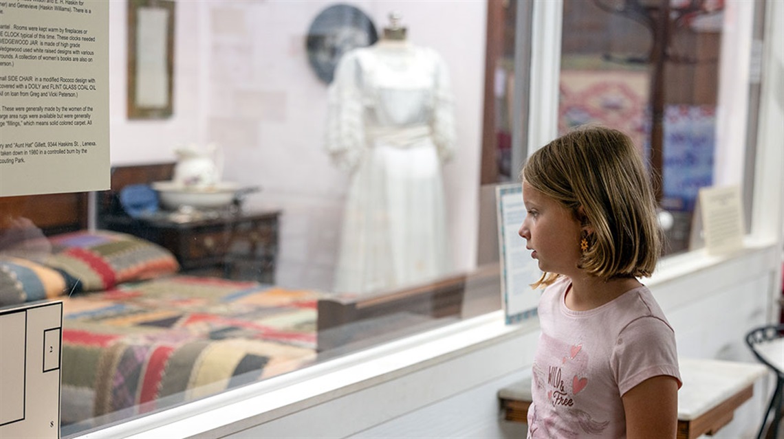 Young girl explores historical exhibit with quilted bed and dress on mannequin