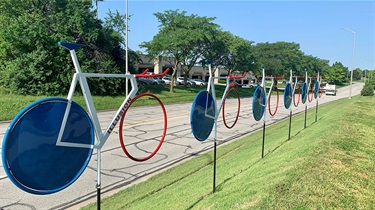 Six red, white and blue painted sculptures in simplified bicycle forms suspended on poles