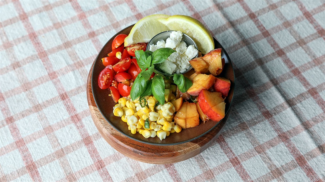 Wooden bowl with composed salad of corn, peaches, tomatoes, basil, goat cheese and lemon wedges on plaid tablecloth