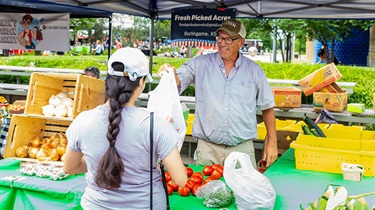 Male vendor hands produce bag to female shopper at Farmers Market booth