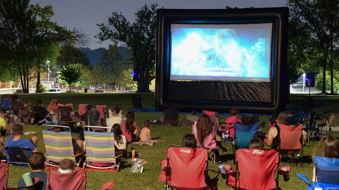 crowd seating in folded chairs watching movie on inflatable screen in park at night