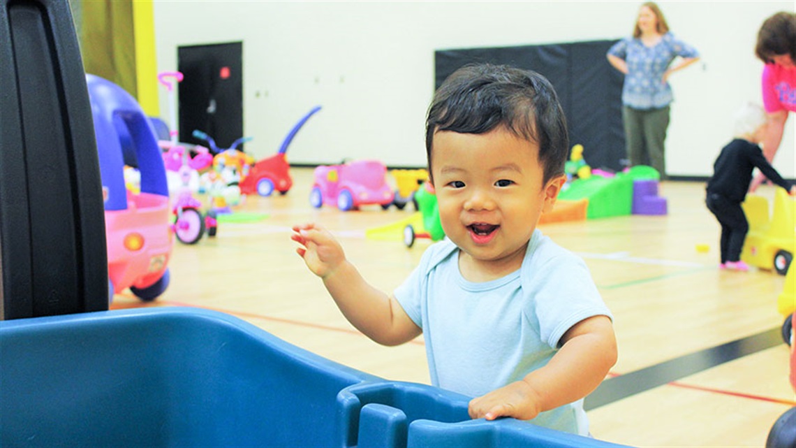 smiling toddler boy holding on to wagon with push toys and cars in the background