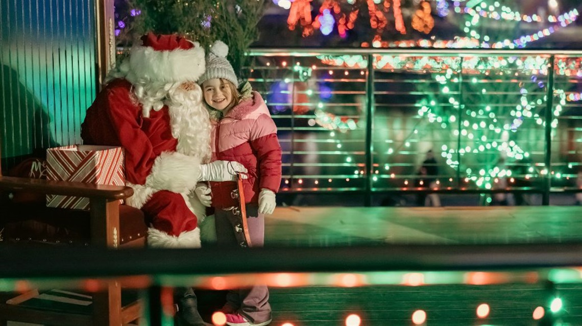Santa Claus hugs smiling young girl in winter attire against park background decorated with holiday lights