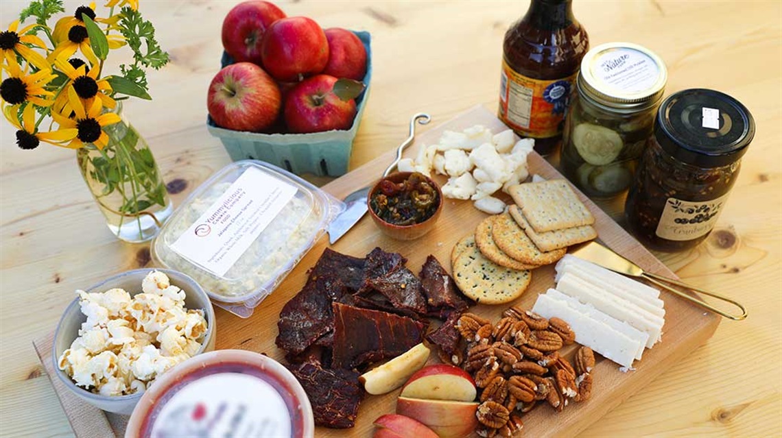 Wood board on table covered with or surrounded by apples, dried meat, cheese, pecans, pickles, barbecue sauce, jam
