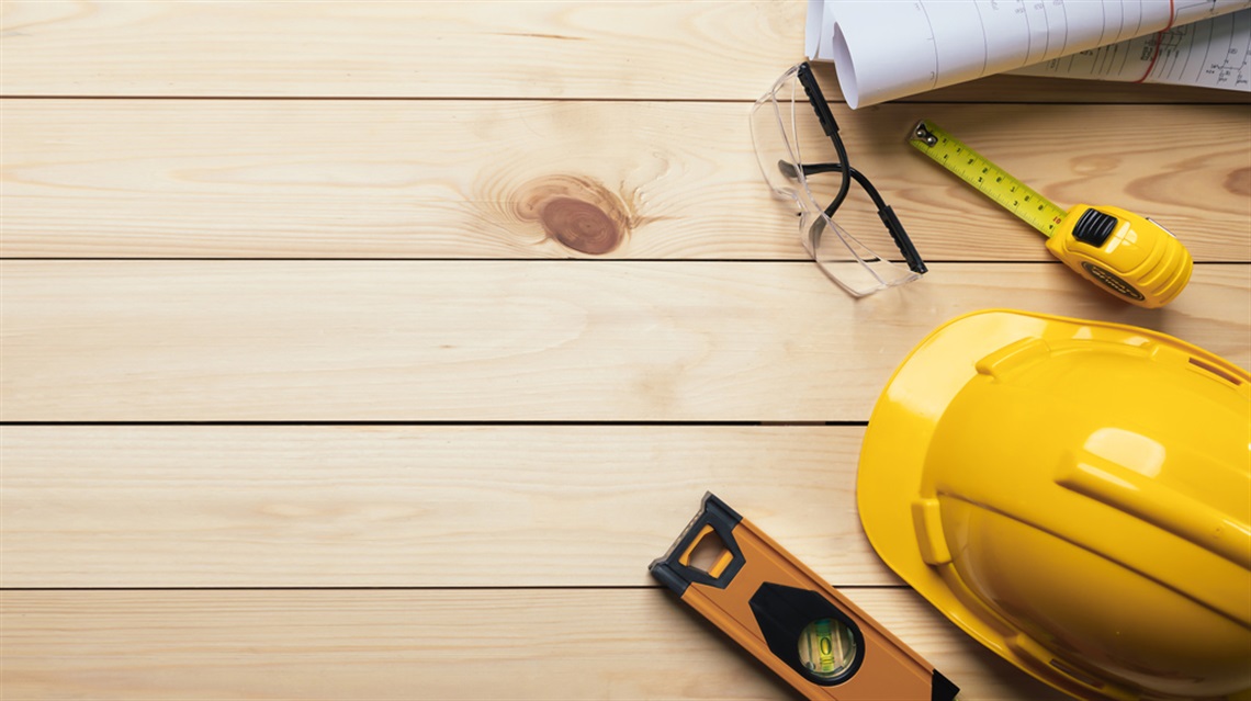 Yellow hardhat, level, tape measure, safety glasses and rolled-up plans laid on wood floor
