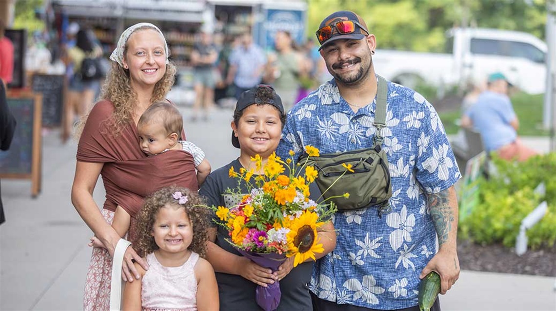 Family of five holding flowers and vegetables at outdoor farmers market