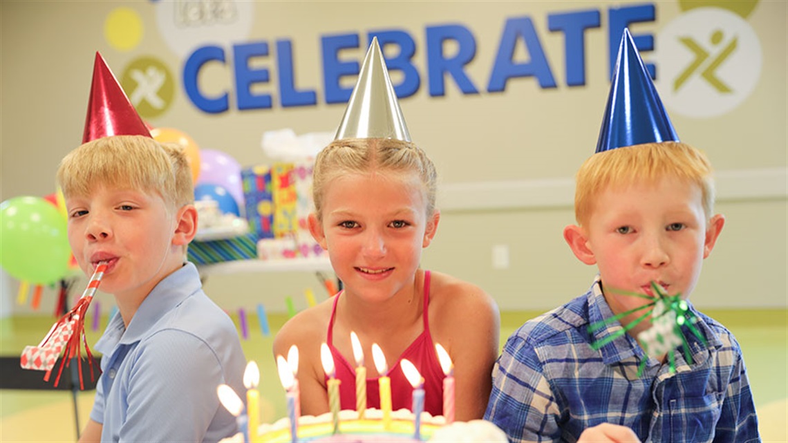 three kids in party hats sitting next to lit birthday cake