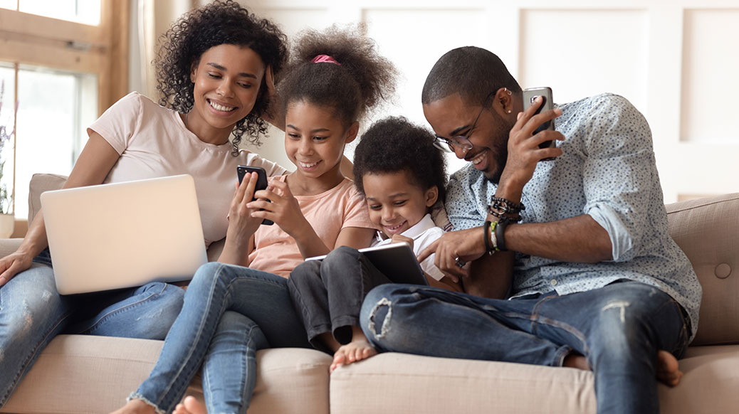 parents and three kids sitting on couch using laptop, tablet, cell phone