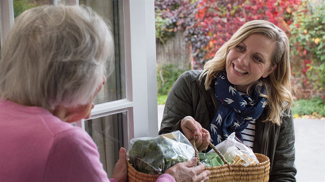 Older woman accepts basket of food from middle-aged woman at front door